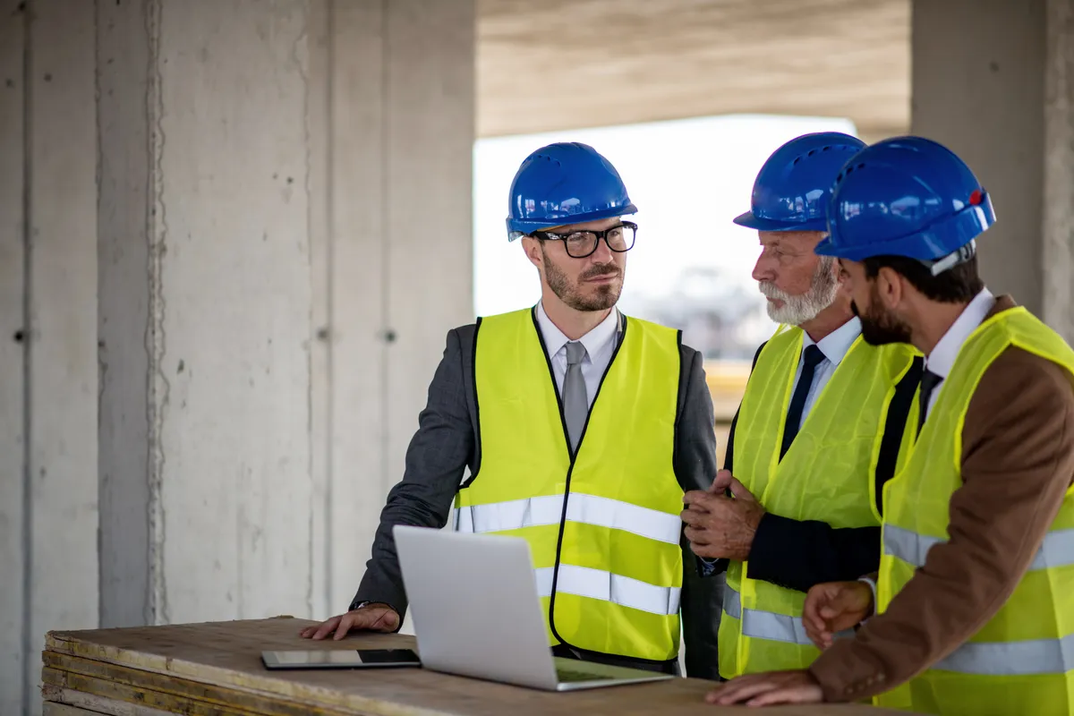 Three engineers in hardhats reviewing a laptop at a concrete construction site